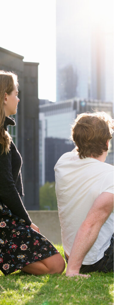 A couple sit in a park looking at the Melbourne skyline.